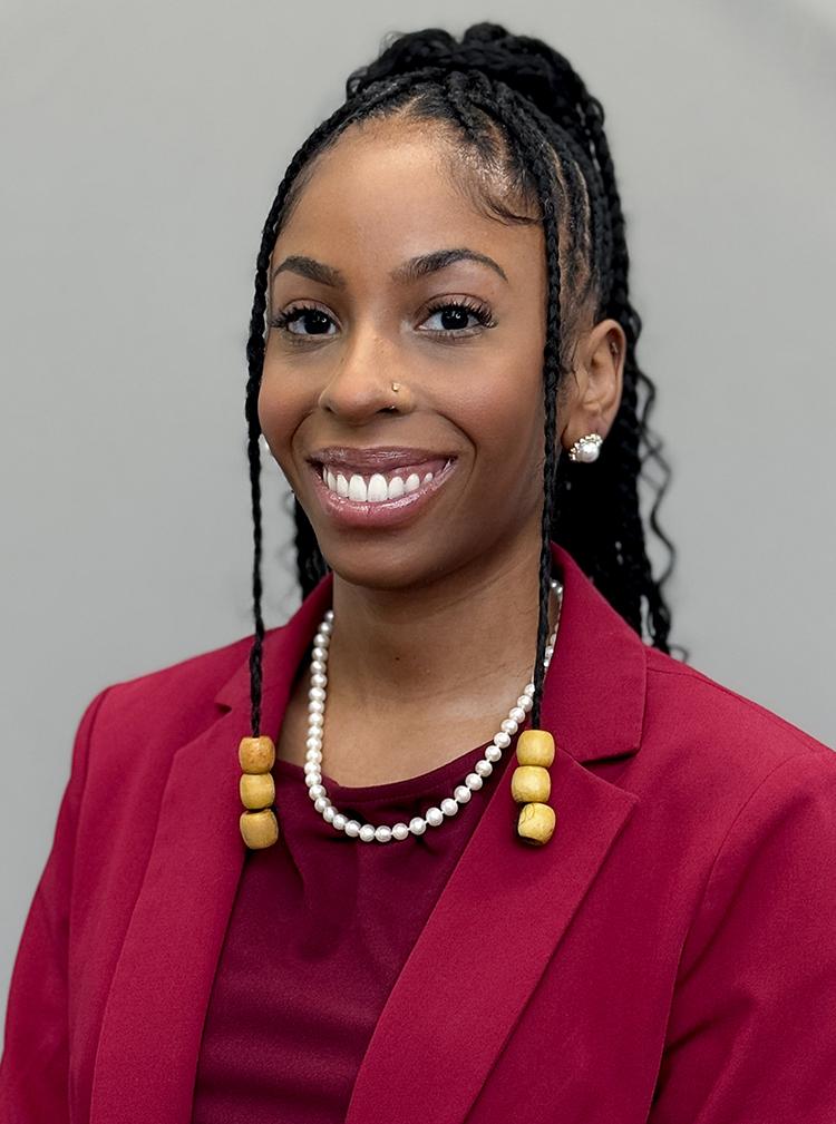 Headshot of Jessica Fuqua, smiling with a red suit on and white pearls. Her hair is braided with beads.
